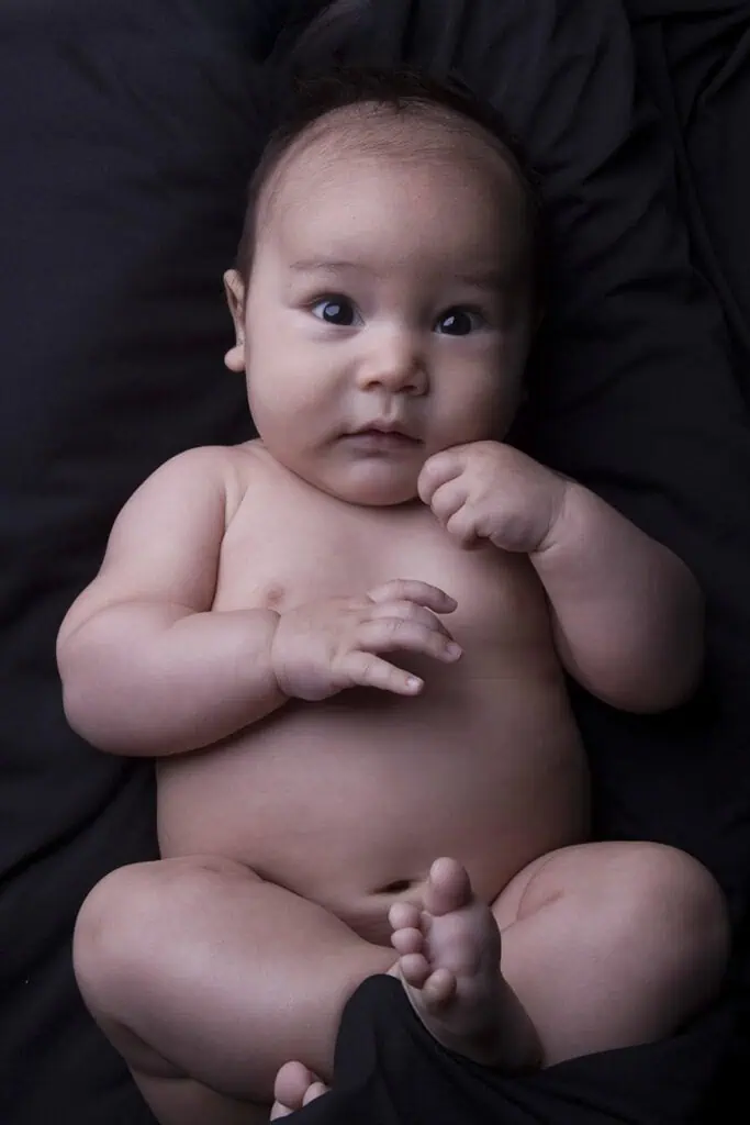 A baby with big brown eyes lies on a black blanket, gazing up with a curious expression, one hand near their mouth and legs slightly bent. - Jodi O Photography