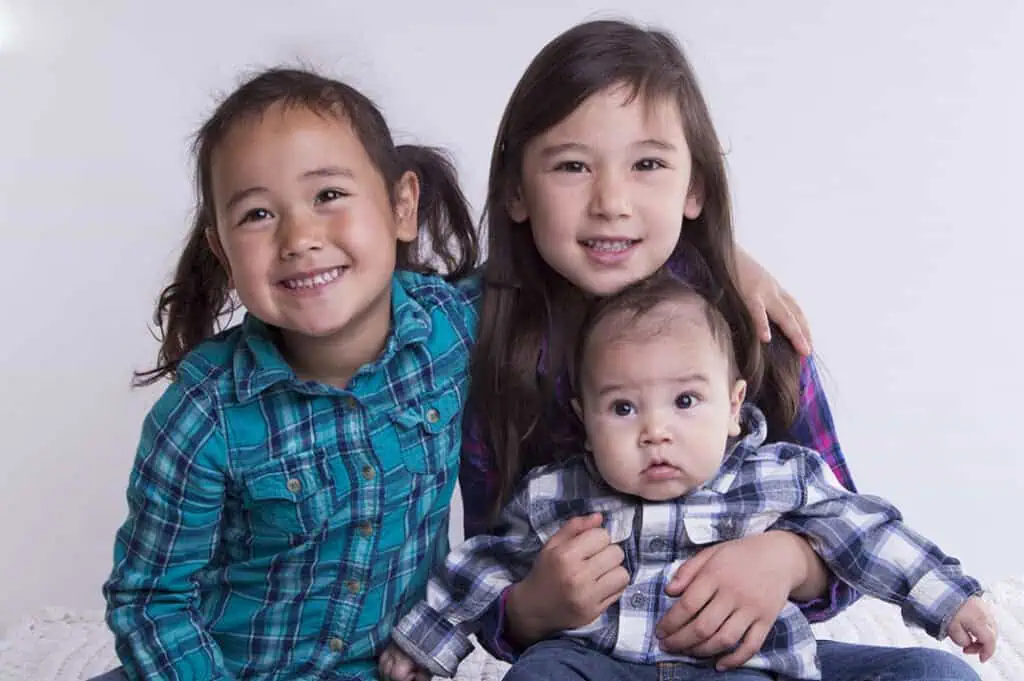 Three young children sit close together and smile. The two girls wear plaid shirts and sit on either side of a baby, who is dressed in a checked shirt and looks slightly to the side. The background is plain white. - Jodi O Photography