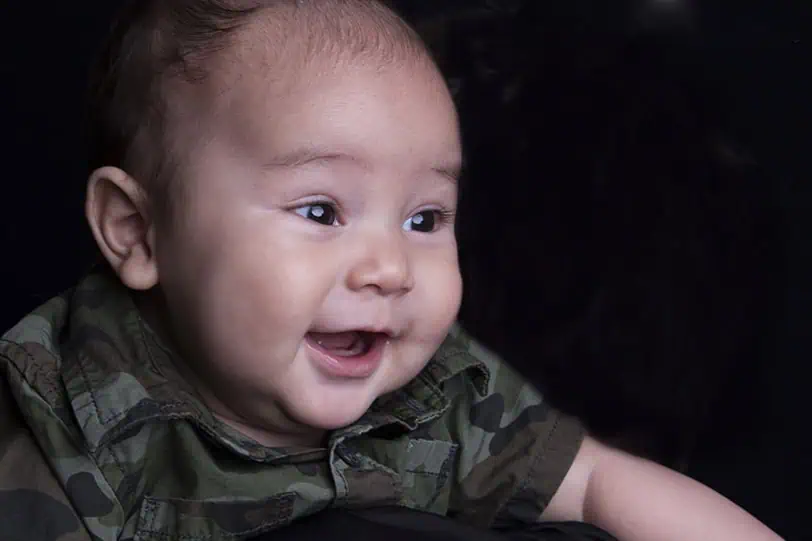 A smiling baby wearing a green camouflage shirt looks off to the side against a dark background. - Jodi O Photography
