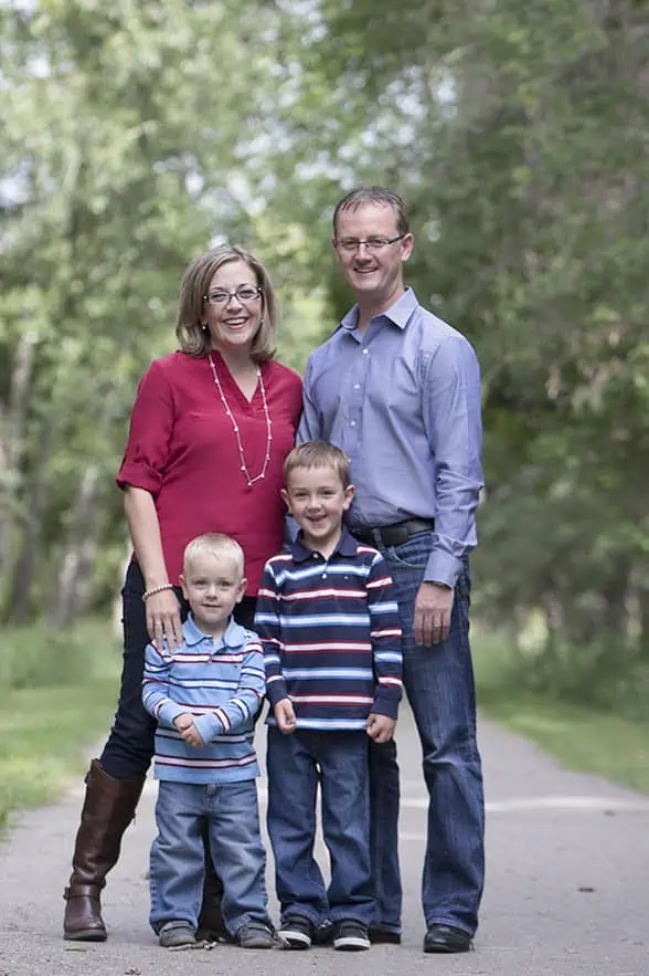 A family of four stands on a tree-lined path, smiling. Two adults stand behind two young boys. All are casually dressed in long-sleeve shirts and jeans. The background is lush and green, suggesting a park or nature trail. - Jodi O Photography