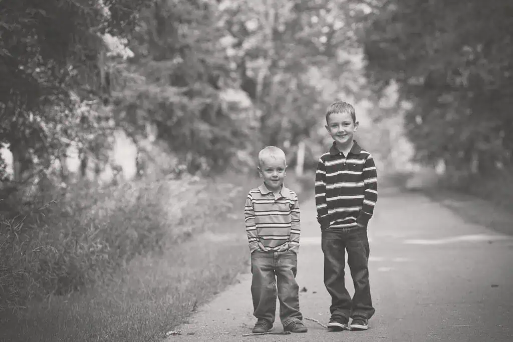 Two young boys stand side by side on a tree-lined path, both wearing striped shirts and jeans. They are smiling at the camera in a black and white outdoor setting. - Jodi O Photography