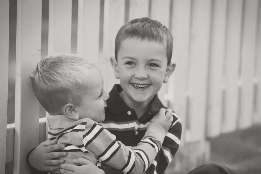 Two young boys sit in front of a white picket fence; the older boy smiles at the camera while hugging the younger boy, who looks up at him. Both wear striped shirts. The scene is in black and white. - Jodi O Photography