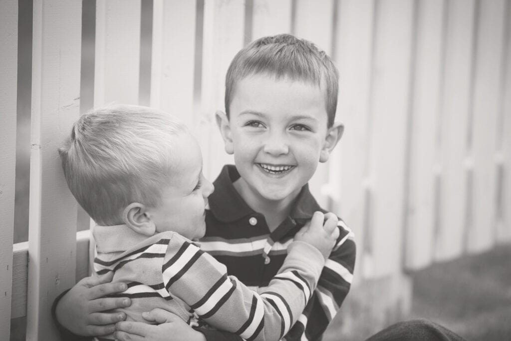 Two young boys sit together by a white wooden fence, both smiling and wearing striped shirts. The older boy hugs the younger, who looks up at him—a heartwarming moment from an outdoor family photo session Calgary families cherish. - Jodi O Photography