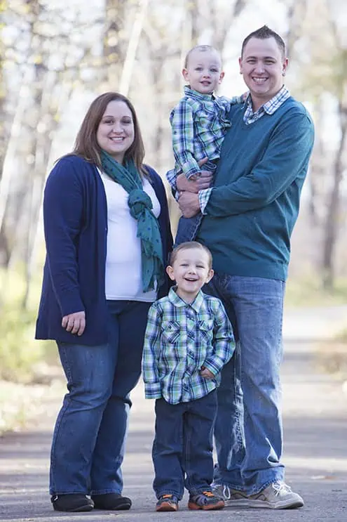 A family of four poses outdoors on a path. Two adults and two young boys, all smiling. The adults and boys wear blue and green outfits. Trees and soft sunlight are in the background. - Jodi O Photography