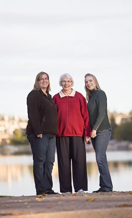 Three women stand close together outdoors, smiling at the camera. The woman in the center has short white hair and wears a red jacket, while the others wear dark jackets. A body of water and trees are visible in the background. - Jodi O Photography