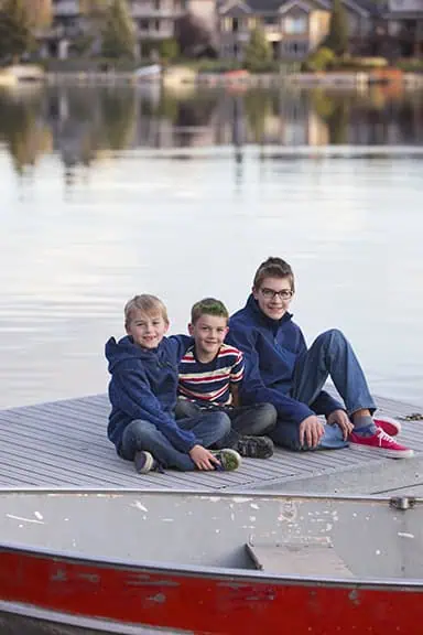 Three boys sit together on a wooden dock by a calm lake, with houses and trees visible in the background. They are smiling and wearing casual clothes, including blue hoodies and jeans. - Jodi O Photography