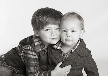 Two young children pose together for a studio portrait. The older child hugs the younger, both wearing plaid shirts and jackets, with neutral expressions. The image is in black and white. - Jodi O Photography