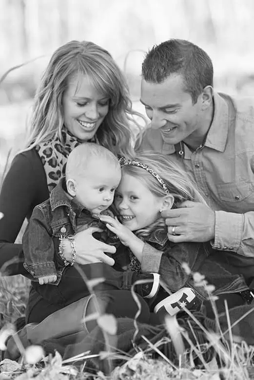 A black and white photo of a smiling family sitting outdoors. Two adults sit behind a young girl and a baby, all cuddling closely together and appearing happy and affectionate. - Jodi O Photography