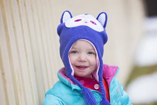A smiling toddler wearing a bright blue jacket and a purple winter hat with animal ears and a white face design stands outdoors near a beige fence. - Jodi O Photography