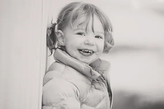 A young child with light hair in pigtails smiles joyfully, showing missing teeth. The child is wearing a puffy jacket and is leaning against a wall. The image is in black and white. - Jodi O Photography