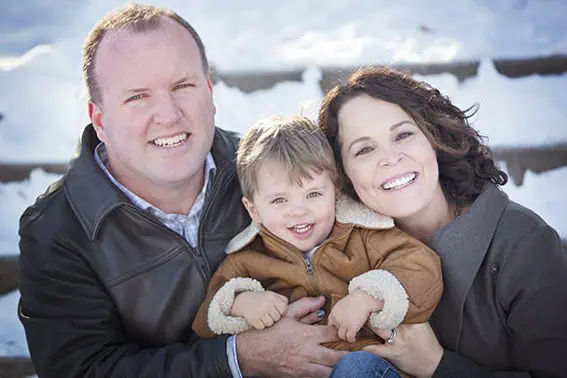 A smiling man, woman, and young child in warm jackets pose closely together outdoors on snowy steps. The family looks happy and enjoys a winter day. - Jodi O Photography