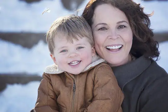 A woman and a young child, both smiling, are outdoors in a snowy setting. The child is wearing a brown jacket, and the woman has dark hair and is wearing a dark coat. They appear happy and are close together. - Jodi O Photography