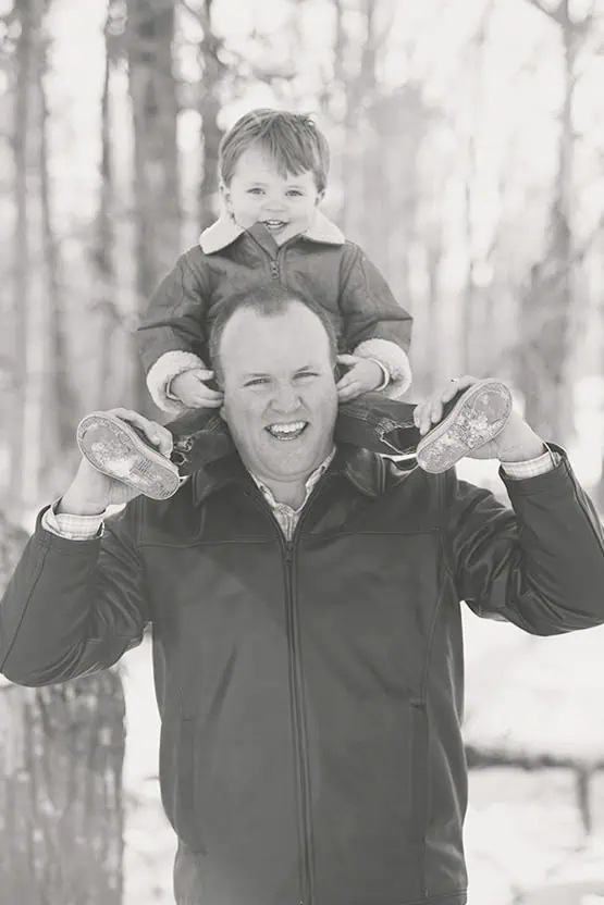 A man in a jacket holds a smiling young child on his shoulders outdoors in a snowy, wooded area. The photo is in black and white. - Jodi O Photography