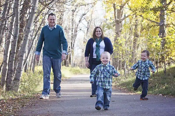 A smiling family of four walks on a tree-lined path in autumn. Two young boys run ahead, while a man and woman walk behind them. Sunlight filters through the yellow and green leaves. - Jodi O Photography