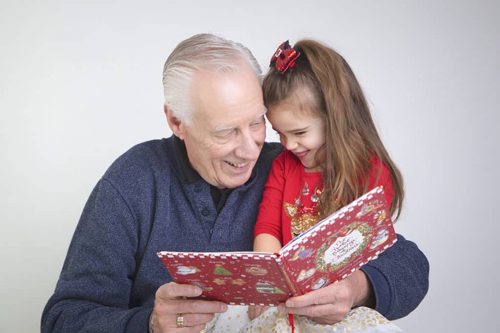 An elderly man and a young girl, both smiling, read a festive red book together during a Studio Family Photos Calgary session. The joyful pair, dressed in vibrant colors, appear engaged and happy as they share this special moment. - Jodi O Photography