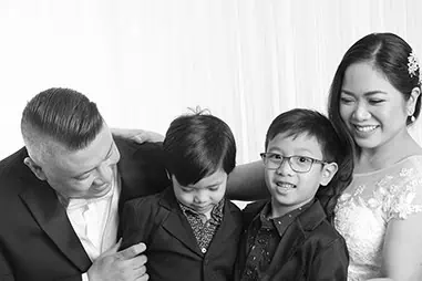 A family of four, dressed formally for their Calgary wedding vow renewals, smiles together. The parents stand on either side of their sons—the younger looks down while the older, wearing glasses, smiles at the camera. - Jodi O Photography