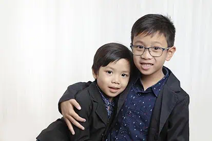 Two young boys in dark blazers pose together in front of a white background. The older boy, wearing glasses, has his arm around the younger boy as they smile gently—capturing the warmth often seen in Calgary family photos. - Jodi O Photography