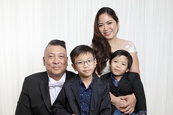 A family of four poses in formal attire against a white backdrop, capturing beautiful Calgary family photos. Two adults and two young boys in suits smile warmly at the camera, dressed for what could be special wedding vow renewals. - Jodi O Photography