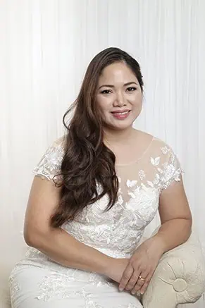 A woman in a lace wedding dress with long, wavy hair sits on a light-colored bench, smiling gently in front of a white, draped background—perfect for Calgary family photos or intimate wedding vow renewals. - Jodi O Photography