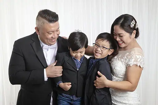 A family of four poses together for family photos in Calgary, smiling. The father and son wear black suits, the younger boy wears a black jacket and jeans, and the mother is in a white lace dress—perfect for wedding vow renewals. A white curtain is in the background. - Jodi O Photography