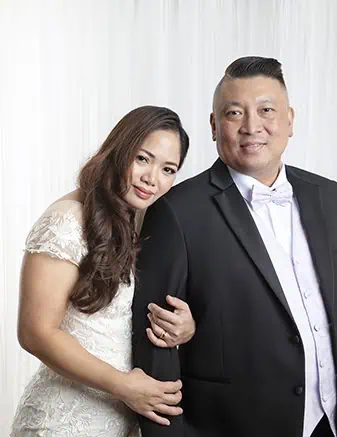 A woman in a white dress holds the arm of a man in a black suit and white bow tie. They stand close together, smiling softly, in front of a white, draped background—perfect for Calgary family photos or wedding vow renewals. - Jodi O Photography