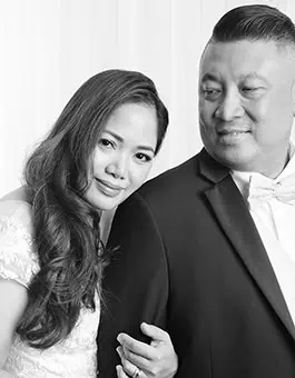 A woman in a white dress leans her head on a man's shoulder, holding his arm. The man wears a suit and bow tie and looks at her, both smiling slightly. Perfect for family photos or wedding vow renewals in Calgary, the photo is black and white with a soft background. - Jodi O Photography