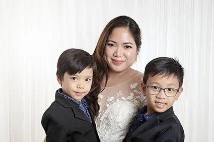 A woman in a white lace dress smiles while posing with two young boys in dark suits against a light, textured background—perfect for capturing heartfelt Calgary family photos or celebrating special wedding vow renewals. - Jodi O Photography