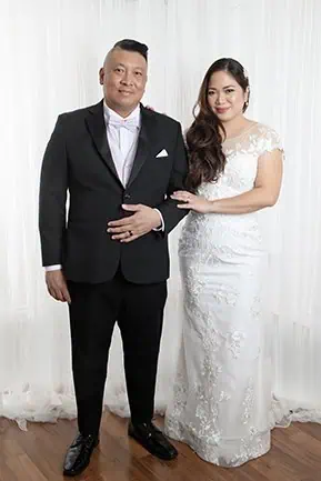 A bride in a white lace gown stands beside a groom in a black suit and bow tie. They pose together, smiling, for wedding vow renewals against a white curtain backdrop on a wooden floor in Calgary. - Jodi O Photography