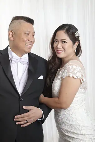 A couple stands together, smiling, in front of a white, draped background. The man wears a black tuxedo and the woman a white lace wedding dress—perfect for capturing family photos or celebrating wedding vow renewals in Calgary. - Jodi O Photography
