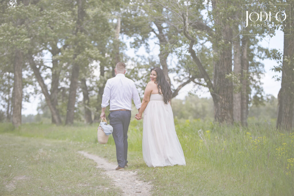 A couple walks hand in hand down a grassy path, the man carrying a picnic basket. The woman, wearing a white dress, looks back and smiles. Tall trees and greenery surround them on a bright, peaceful day. - Jodi O Photography