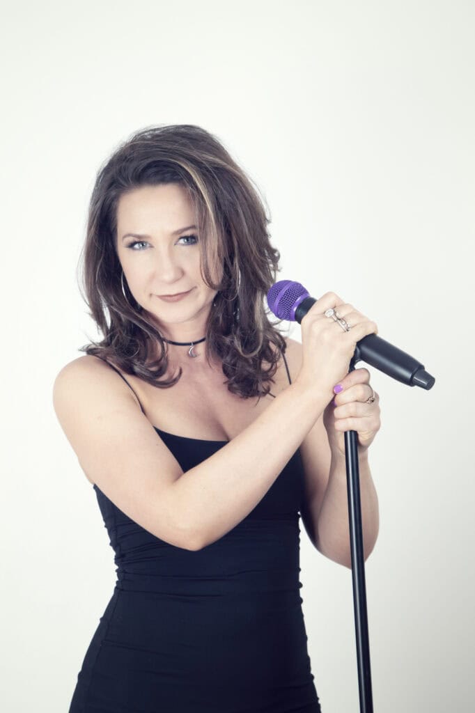 A woman with wavy brown hair wearing a black sleeveless dress holds a purple microphone on a stand, smiling slightly as if ready to record in a music studio, standing against a plain white background. - Jodi O Photography