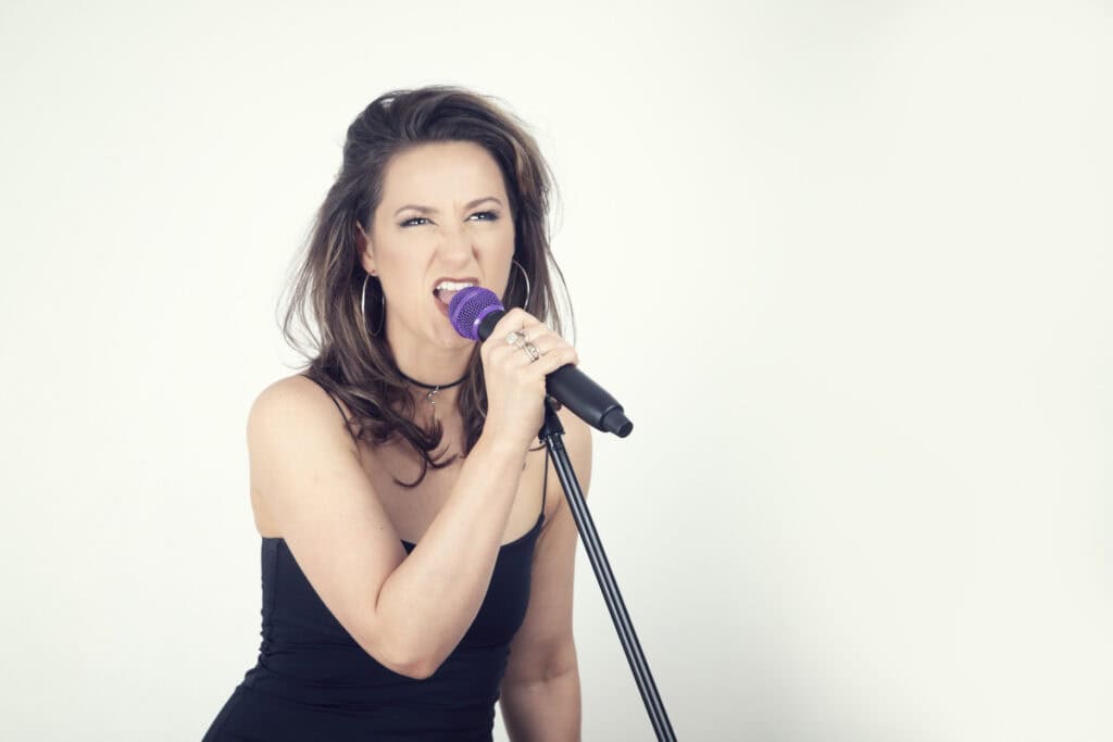 A woman with long brown hair in a black strapless dress passionately sings into a purple microphone, exuding Rock Star Energy with an expressive, intense look, as if performing her heart out in a music studio against a plain white background. - Jodi O Photography