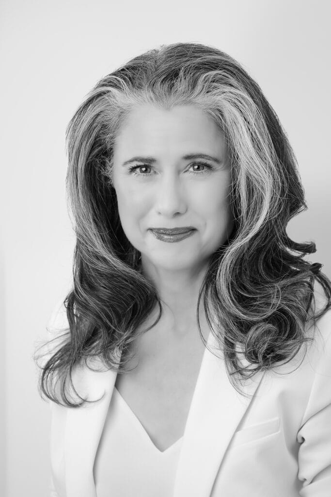 Black and white portrait of a confident woman with long, wavy, dark hair streaked with gray, wearing a light-colored blazer and top, smiling softly at the camera against a plain background. - Jodi O Photography