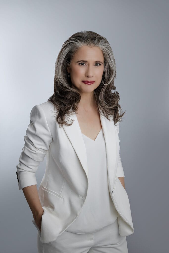 A professional woman with long, wavy gray hair wearing a white blazer and matching top and pants stands confidently with her hands in her pockets against a plain gray background. - Jodi O Photography