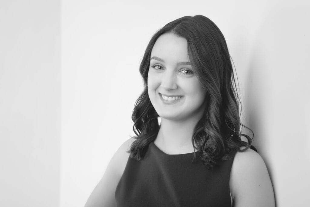 A wedding planner with wavy dark hair, wearing a sleeveless dark top, smiles while leaning against a white wall in a black and white portrait, perfectly capturing her passion for building a brand. - Jodi O Photography