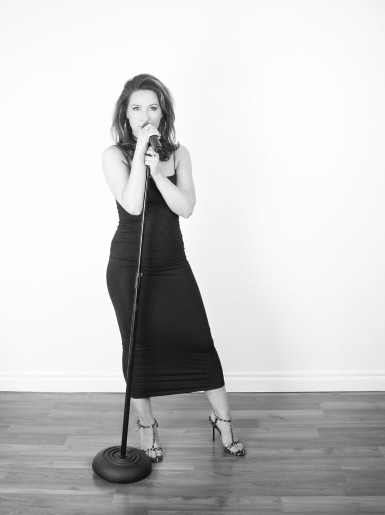 A woman in a fitted, sleeveless black dress stands on a wooden floor in a music studio, holding a microphone on a stand and singing. The background is plain white, and the photo is in black and white. - Jodi O Photography