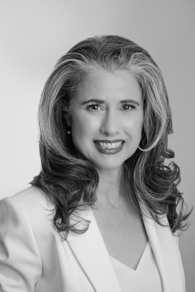 Black and white portrait of an authentic, confident woman with wavy, shoulder-length hair, smiling and wearing a light-colored blazer over a top, against a plain background. - Jodi O Photography