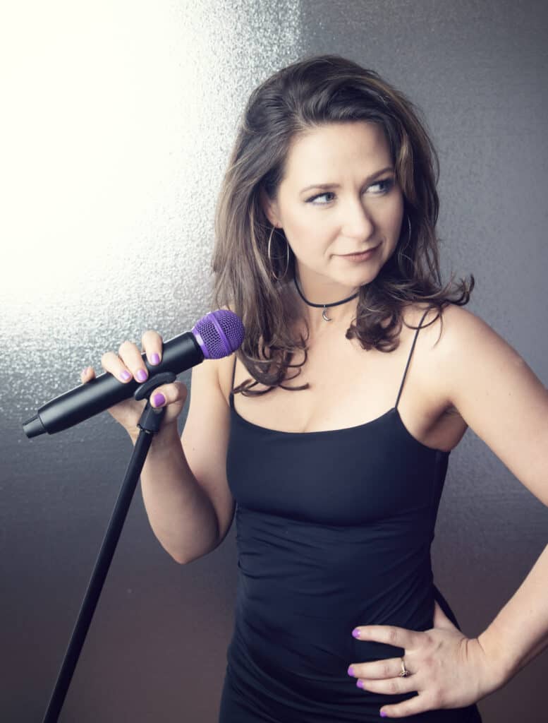 A woman in a black dress holds a purple microphone on a stand, looking to the side with a confident expression, as if ready to step into a music studio for her next recording session. - Jodi O Photography