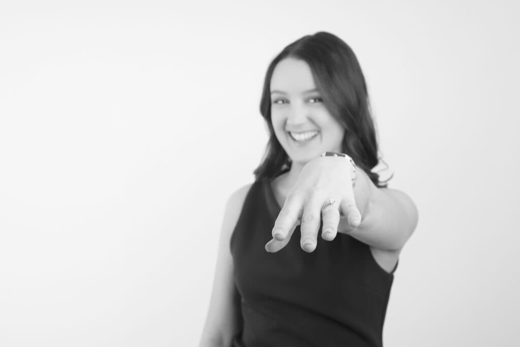 A smiling woman with shoulder-length hair extends her left hand forward, displaying a ring on her finger—a classic black and white photo capturing the excitement of wedding planning, set against a plain light background. - Jodi O Photography
