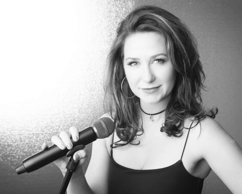 A woman with wavy hair, wearing a sleeveless top and hoop earrings, smiles while holding a microphone in what appears to be a studio, set against a textured, light background. The image is in black and white. - Jodi O Photography