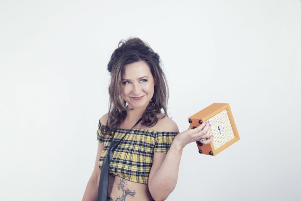 A smiling woman with wavy brown hair, wearing a yellow plaid off-shoulder top and a necktie, holds a small orange guitar amplifier in her left hand, exuding Rock Star Energy in this bright studio shot against a plain white background. - Jodi O Photography
