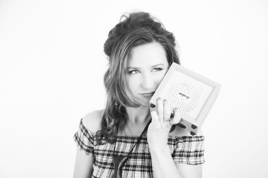 A woman with wavy hair in an off-the-shoulder plaid top holds a small square speaker to her face, looking thoughtfully to the side. Captured in black and white, she exudes effortless Rock Star Energy against a plain studio backdrop. - Jodi O Photography