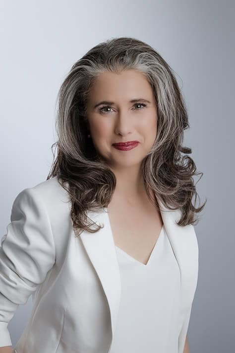 A confident woman with long, wavy gray hair wearing a white blazer and top smiles softly, posing authentically against a plain light gray background. - Jodi O Photography