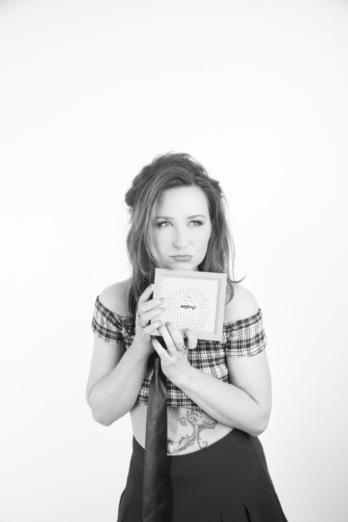 A woman with long hair, wearing an off-shoulder plaid top, holds a small patterned box close to her face. With a thoughtful expression and a visible tattoo, she looks like she’s lost in thought at a music studio. The image is in black and white. - Jodi O Photography