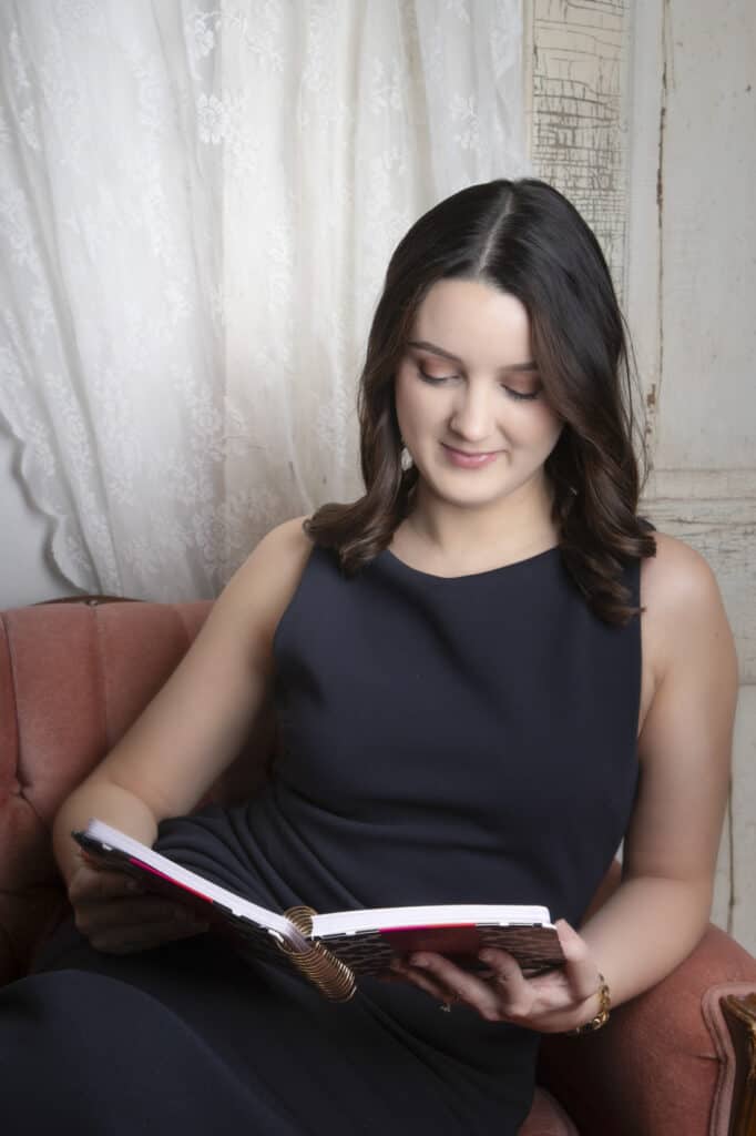 A wedding planner with shoulder-length dark hair sits on a pink armchair, wearing a sleeveless black dress and reading an open notebook. White lace curtains and a distressed wooden wall are in the background. - Jodi O Photography