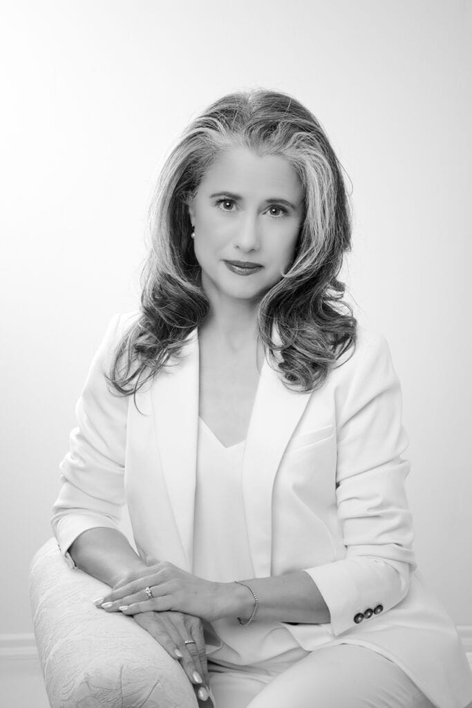 A grayscale portrait of a confident woman with long, wavy hair wearing a light-colored suit. She is seated, looking at the camera with a gentle expression, hands resting on an armrest. The background is plain and bright. - Jodi O Photography