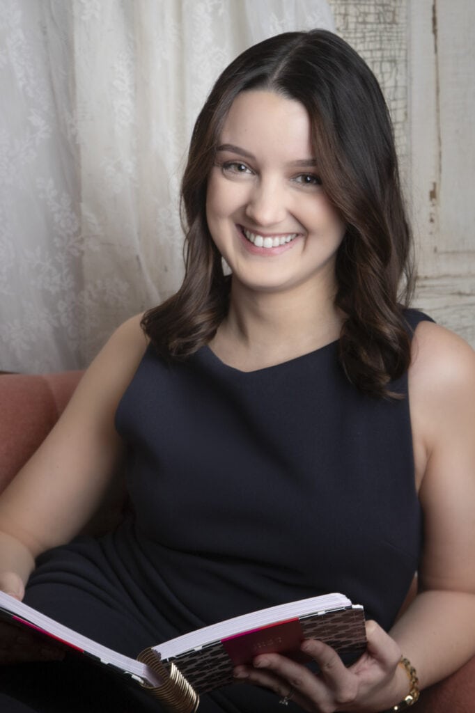 A wedding planner with shoulder-length brown hair, wearing a sleeveless dark dress, sits on a pink sofa smiling and holding an open notebook in her lap. A white lace curtain and rustic wooden panel are in the background. - Jodi O Photography