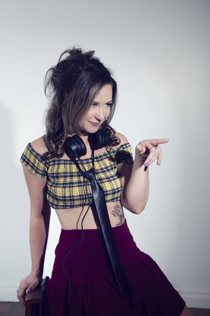 In a studio setting, a woman with wavy brown hair wears a plaid crop top, burgundy skirt, and black necktie. With headphones around her neck and a visible side tattoo, she points to the side and smiles against a plain background. - Jodi O Photography