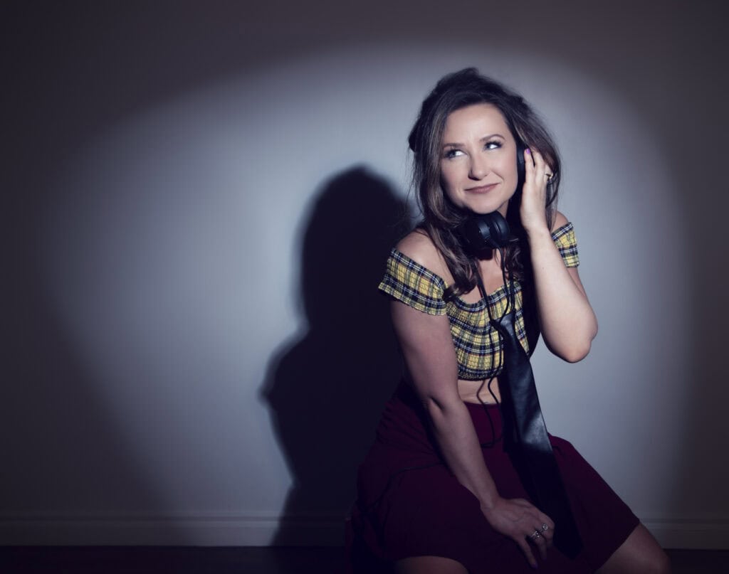 A woman with long brown hair, wearing a yellow plaid off-the-shoulder top and a dark skirt, sits against a wall with headphones around her neck. She smiles softly, one hand touching the headphones, looking like she's just left the music studio. - Jodi O Photography