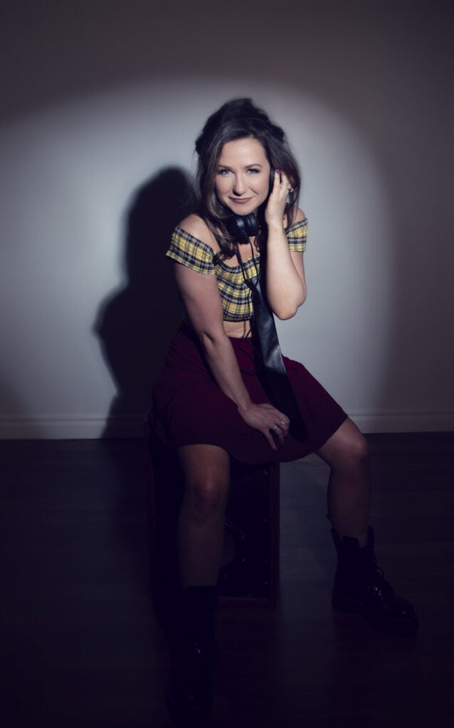 A woman sits on a stool in a dimly lit music studio, smiling at the camera. She wears a yellow plaid crop top, red skirt, black boots, and a black tie. One hand touches her hair as Rock Star Energy radiates under the spotlight against a plain wall. - Jodi O Photography